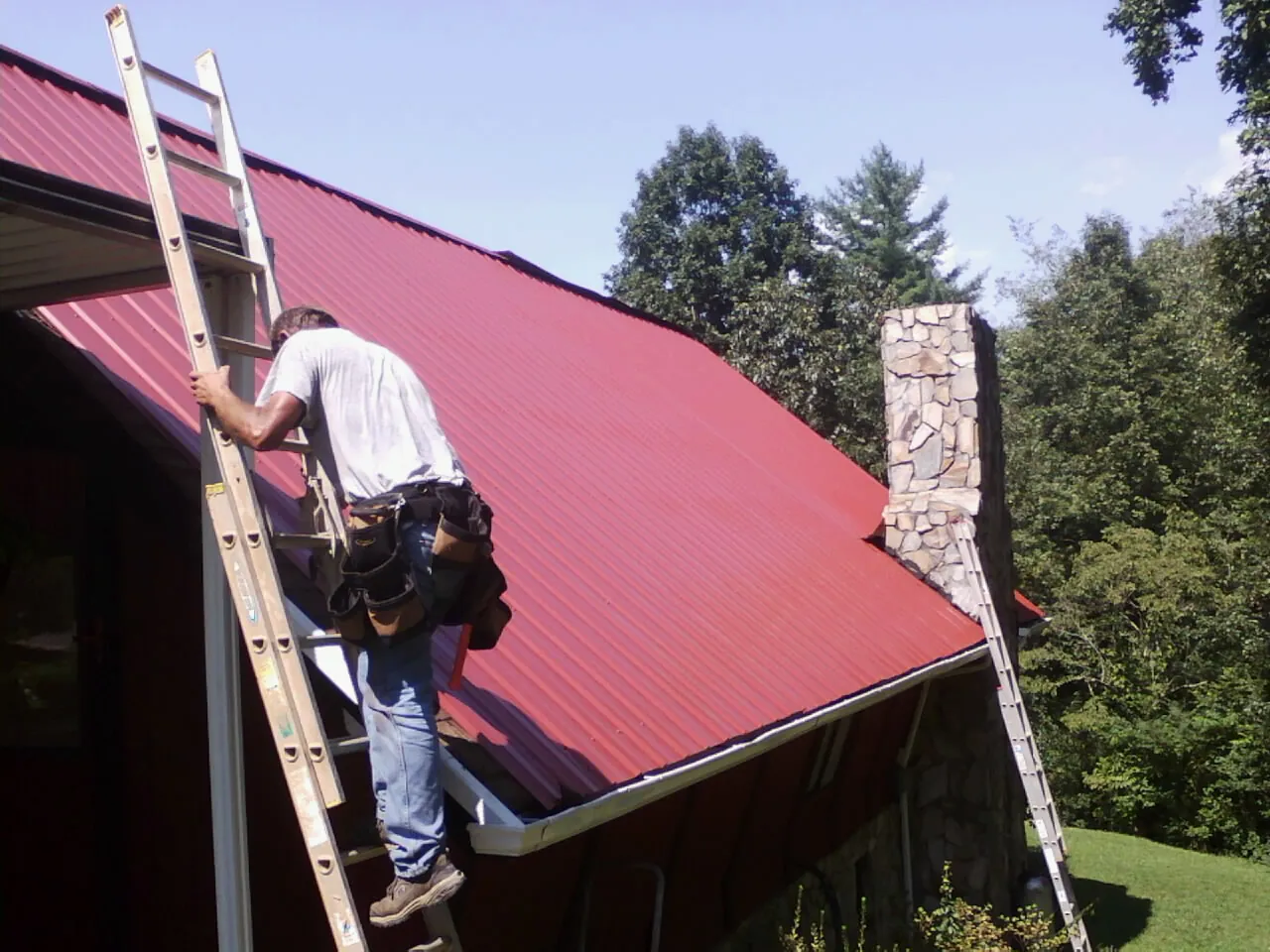 Roofer on a ladder beside a red metal roof and stone chimney — Soffit & Fascia Repair by Roofing Brothers in Cherry Point